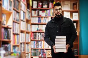 studente arabo alto e intelligente, indossa una giacca di jeans nera e occhiali da vista, in biblioteca con una pila di libri. foto