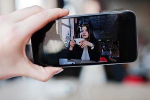 foto ravvicinata di telefoni cellulari con schermo con ragazza bruna seduta al bar con una tazza di cappuccino, ascoltando musica con le cuffie.