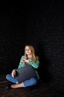 ragazza bionda alla moda in giacca e jeans con il computer portatile contro il muro nero di mattoni in studio. foto