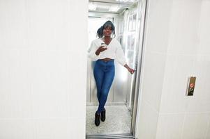 eleganti donne afroamericane in camicetta bianca e jeans blu poste in ascensore con il telefono cellulare a portata di mano. foto