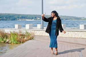 modello afroamericano dalla pelle scura plus size posato con un vestito blu lucido e una pelliccia nera contro il lato del mare che fa selfie sul telefono. foto