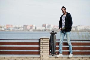 street style uomo arabo in occhiali con longboard poste contro il fiume. foto