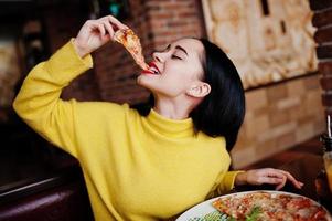 divertente ragazza bruna in maglione giallo che mangia pizza al ristorante. foto