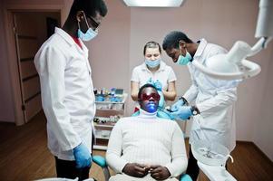 team multirazziale di medici dentisti. paziente uomo afroamericano con occhiali protettivi uv. i suoi denti sono stati trattati con l'aiuto di una lampada a polimerizzazione uv dentale e uno specchio dentale. foto