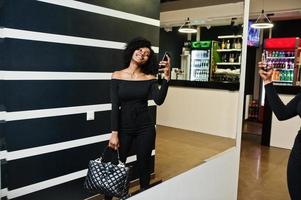 alla moda giovane bella donna afroamericana con acconciatura afro e hangbag in posa usura in nero elegante. fare foto allo specchio per telefono.