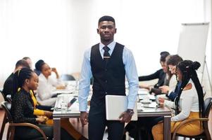 volto di un bell'uomo d'affari africano, con in mano il laptop sullo sfondo della riunione del team multirazziale della gente d'affari, seduto al tavolo dell'ufficio. foto