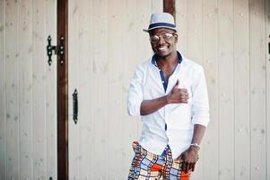 elegante uomo afroamericano in camicia bianca e pantaloni colorati con cappello e occhiali in posa all'aperto. ragazzo nero modello alla moda. foto