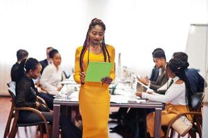 volto di una bella donna d'affari africana in abito giallo, che tiene appunti sullo sfondo della riunione del team multirazziale di uomini d'affari, seduto al tavolo dell'ufficio. foto
