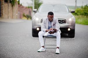 uomo afroamericano ricco ed elegante in giacca e pantaloni bianchi, occhiali con rivista seduto su una sedia contro l'auto suv. foto