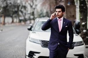 elegante modello di uomo macho indiano su abito e cravatta rosa in posa contro un'auto bianca e parlando al telefono. foto