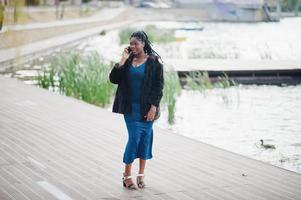 modello afroamericano dalla pelle scura plus size posato con un abito blu lucido e una pelliccia nera contro il lago e parlando al telefono cellulare. foto