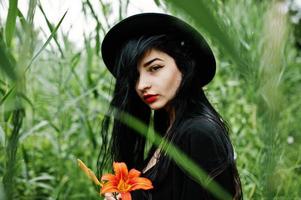 ragazza sensuale tutta in nero, labbra rosse e cappello. goth donna drammatica tenere fiore di giglio arancione su canna comune. foto