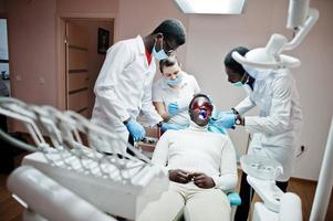 team multirazziale di medici dentisti. paziente uomo afroamericano con occhiali protettivi uv. i suoi denti sono stati trattati con l'aiuto di una lampada a polimerizzazione uv dentale e uno specchio dentale. foto