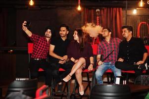 gruppo di amici indiani seduti al lounge bar, divertendosi e riposando, fumando narghilè e guardando sui telefoni cellulari per fare selfie. foto