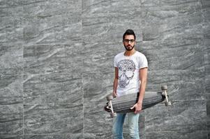 street style uomo arabo in occhiali con longboard poste contro il muro grigio. foto