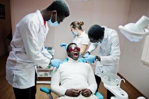 team multirazziale di medici dentisti. paziente uomo afroamericano con occhiali protettivi uv. i suoi denti sono stati trattati con l'aiuto di una lampada a polimerizzazione uv dentale e uno specchio dentale. foto