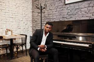 forte potente uomo afroamericano in abito nero seduto contro il pianoforte. foto