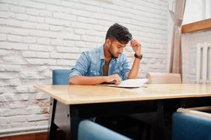 ritratto di bel successo barbuto del sud asiatico, giovane libero professionista indiano in camicia di jeans blu seduto in un bar e menu di lettura. foto