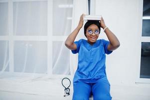 ritratto di felice femmina afroamericano giovane medico pediatra in uniforme blu cappotto e stetoscopio con libri a portata di mano. sanitario, medico, specialista in medicina - concetto. foto