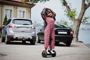 bella donna afroamericana utilizzando segway o hoverboard. ragazza nera su scooter elettrico autobilanciante a doppia ruota contro il parcheggio. foto