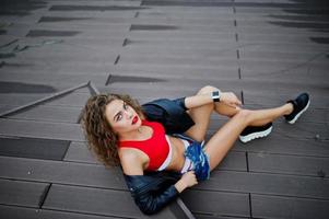 ragazza modello riccia sexy in top rosso, pantaloncini di jeans, giacca di pelle e scarpe da ginnastica poste sul pavimento. foto