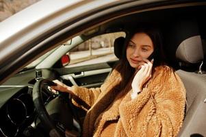 giovane donna d'affari in pelliccia seduta al volante di un'auto e parlare al cellulare. foto