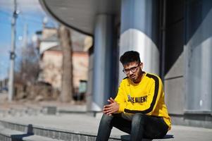 uomo asiatico in occhiali e felpa gialla seduto sulle scale contro un nuovo edificio moderno. foto