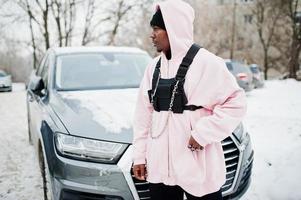l'uomo afroamericano alla moda in stile urbano in felpa con cappuccio rosa ha posato contro l'auto suv in inverno. ragazzo afro rapper. foto
