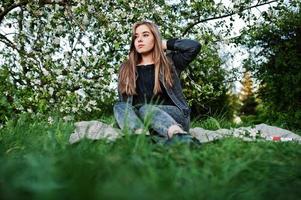 giovane ragazza bruna in jeans seduto sul plaid contro l'albero del fiore di primavera. foto