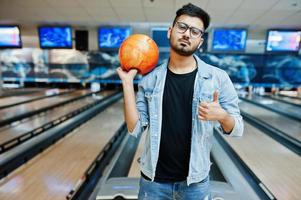uomo asiatico con barba elegante in giacca di jeans e occhiali in piedi alla pista da bowling con palla a portata di mano. foto