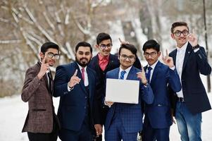 un gruppo di sei uomini d'affari indiani in giacca e cravatta poste all'aperto in una giornata invernale in Europa, guardando sul laptop e mostrando le dita in alto. foto