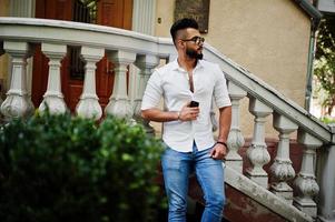 elegante uomo arabo alto modello in camicia bianca, jeans e occhiali da sole posato in una strada della città. barba attraente ragazzo arabo. foto