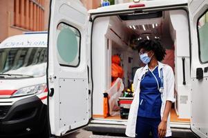 paramedico femminile afroamericano in maschera medica protettiva per il viso in piedi davanti all'auto ambulanza. foto