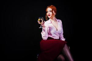 splendida ragazza dai capelli rossi in camicetta rosa e gonna rossa con un bicchiere di vino a portata di mano isolato su nero. foto