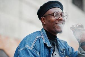 primo piano ritratto di uomo afroamericano in giacca di jeans, berretto e occhiali, sigaro fumante. foto