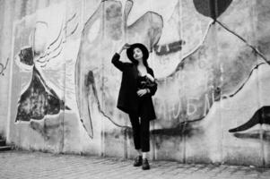 ragazza sensuale tutta in nero, labbra rosse e cappello. la donna drammatica goth tiene il fiore del crisantemo bianco contro il muro dei graffiti. ritratto in bianco e nero. foto
