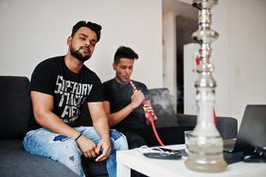due uomini indiani belli e alla moda che si rilassano in camera e fumano narghilè. foto