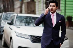 elegante modello di uomo macho indiano su abito e cravatta rosa in posa contro un'auto bianca e parlando al telefono. foto