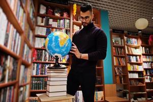 alto e intelligente studente arabo, indossa un dolcevita viola e occhiali da vista, in biblioteca tenendo il globo terrestre a portata di mano. foto