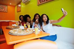 quattro giovani ragazze africane in un ristorante dai colori vivaci che mangiano pizza, si divertono insieme e fanno selfie al telefono. foto