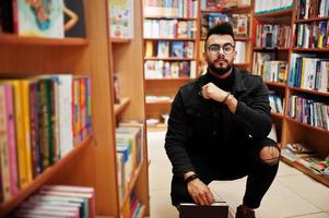 studente arabo alto e intelligente, indossa una giacca di jeans nera e occhiali da vista, in biblioteca con un libro a portata di mano. foto