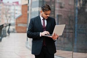 elegante uomo d'affari indiano in abbigliamento formale con laptop sulle mani in piedi contro le finestre nel business center. foto