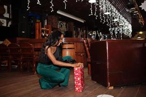 bellezza sottile modello afroamericano indossare su camicetta nera e pantaloni verdi gambe lunghe in posa contro decorazioni natalizie e tenere una confezione regalo per le celebrazioni del nuovo anno. foto