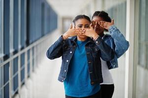 due amiche africane in giacca di jeans coprirono la bocca e gli occhi con le mani al coperto insieme. foto