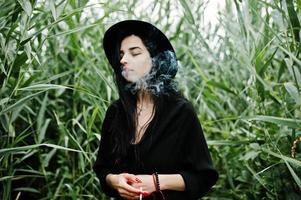 sensuale fumatrice tutta in nero, labbra rosse e cappello. donna drammatica goth che fuma sulla canna comune. foto