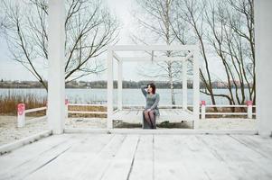 ritratto di ragazza bruna in abito grigio seduto a una costruzione in legno bianco. foto