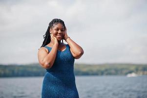 modello afroamericano dalla pelle scura plus size posato in un abito blu lucido contro il lato del mare. foto