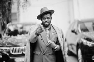 elegante modello uomo afroamericano in cappotto grigio, giacca cravatta e cappello rosso contro due suv nere d'affari e mostra il segno bang bang, tema gangster. foto in bianco e nero.