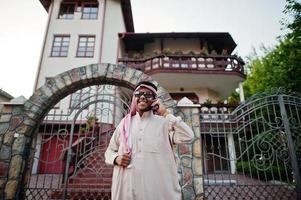 ricco uomo d'affari arabo mediorientale con occhiali da sole in posa per strada contro il palazzo, parlando al telefono cellulare. foto