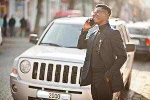 l'uomo afroamericano dall'aspetto sorprendente indossa un blazer blu con spilla, dolcevita nero e occhiali in posa in strada. ragazzo nero alla moda che parla al telefono contro l'auto. foto